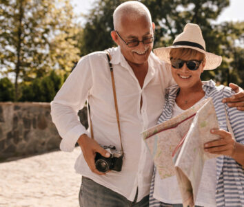 Cheerful man with grey hair in light shirt and jeans with camera smiling and looking at map with blonde lady in hat and blue outfit in park.